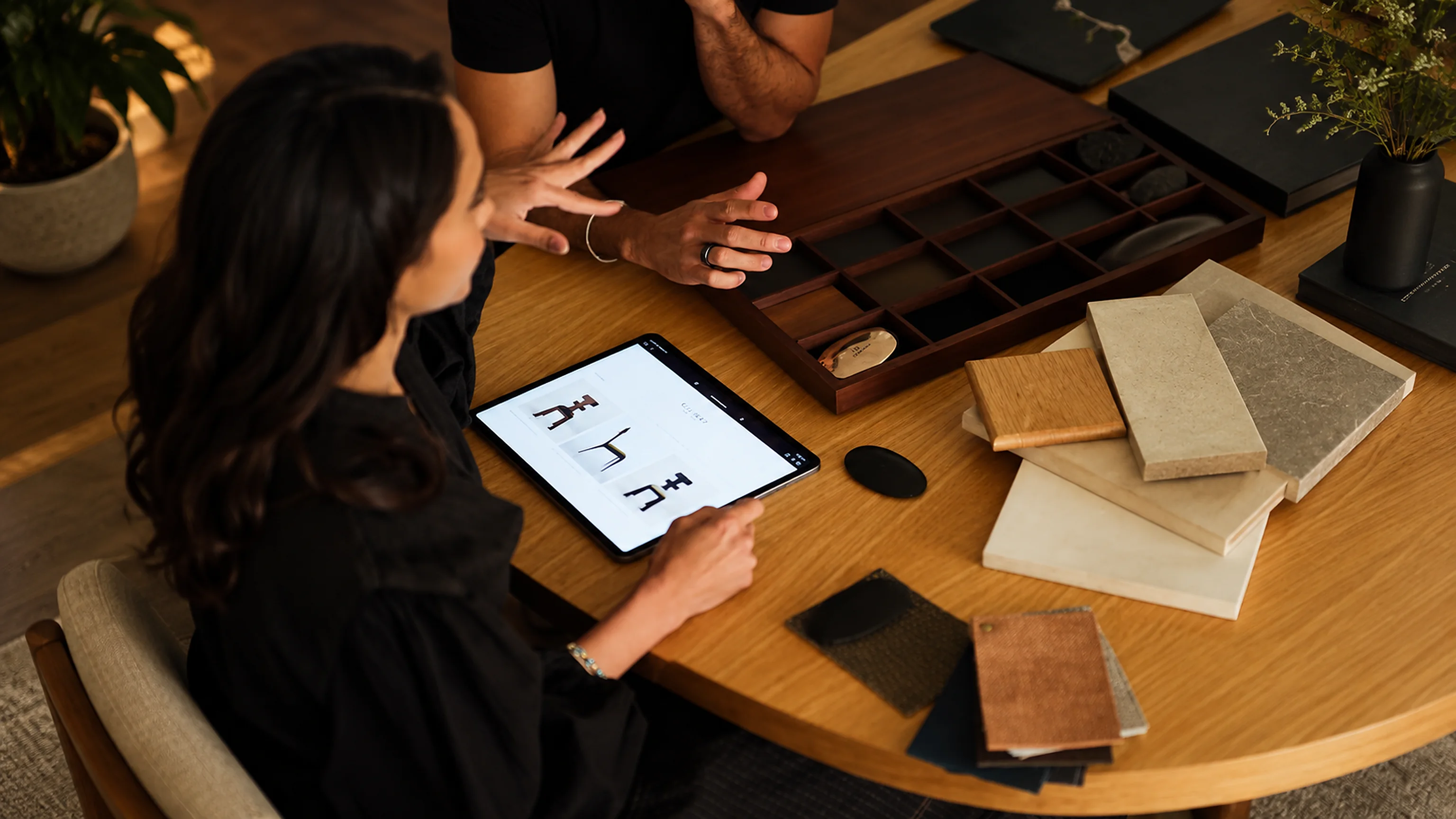 Two people at a round table looking at a project on a tablet, among sample trays and notebooks