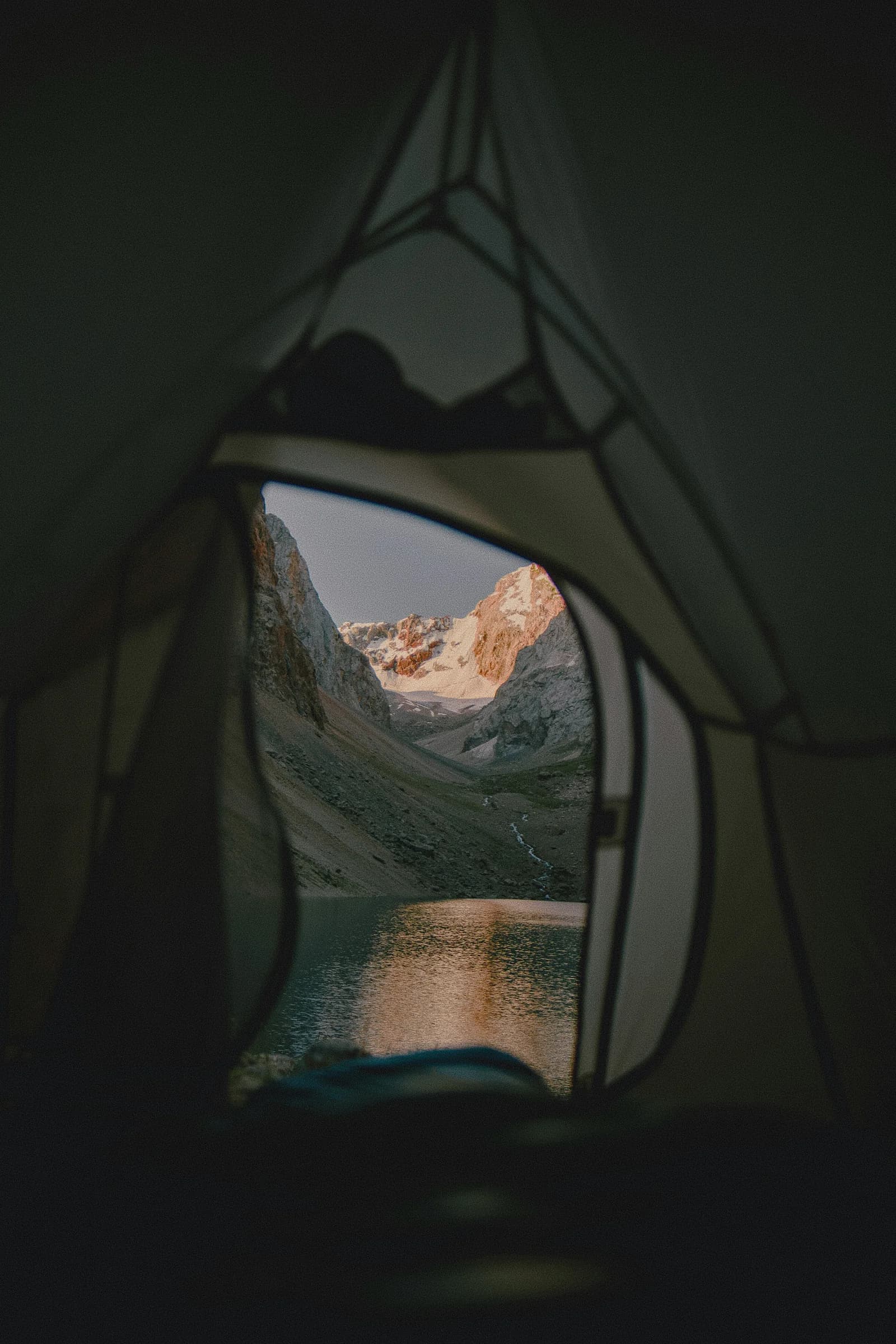 L’apertura di una tenda inquadra una montagna innevata e l’acqua ferma di un lago