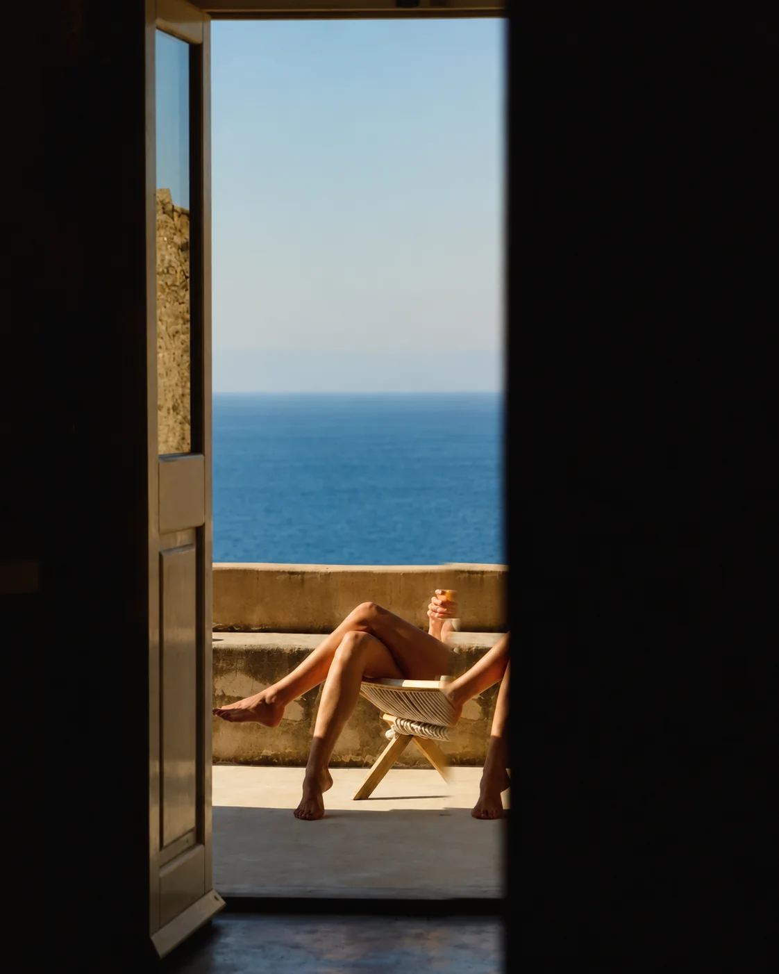 Una porta scura inquadra una terrazza sul mare, con una persona seduta al sole