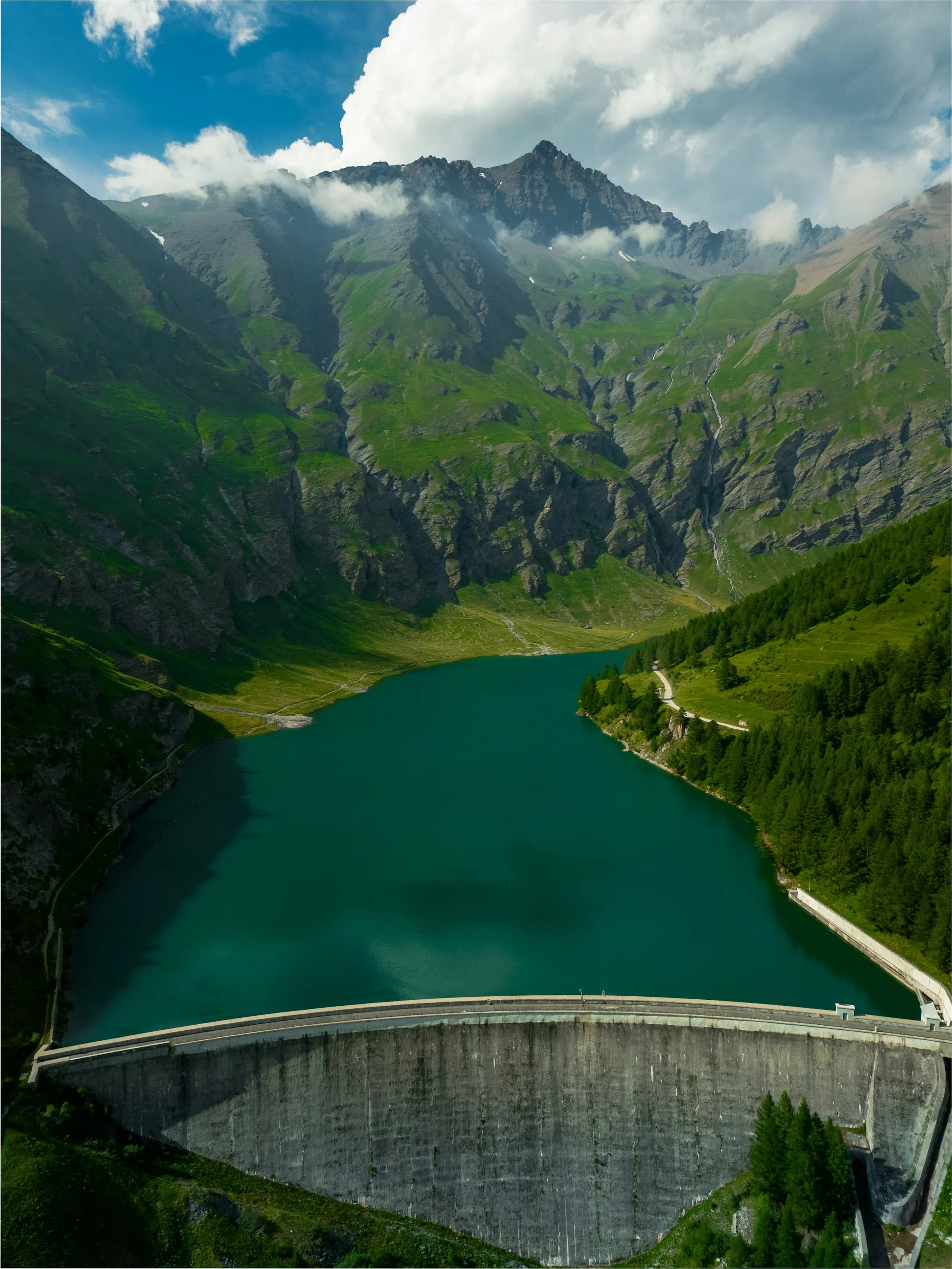 Il lago di Rochemolles visto dall’alto: l’acqua verde scuro chiusa dalla diga di cemento, i pascoli e le pareti di roccia della valle sotto le nuvole
