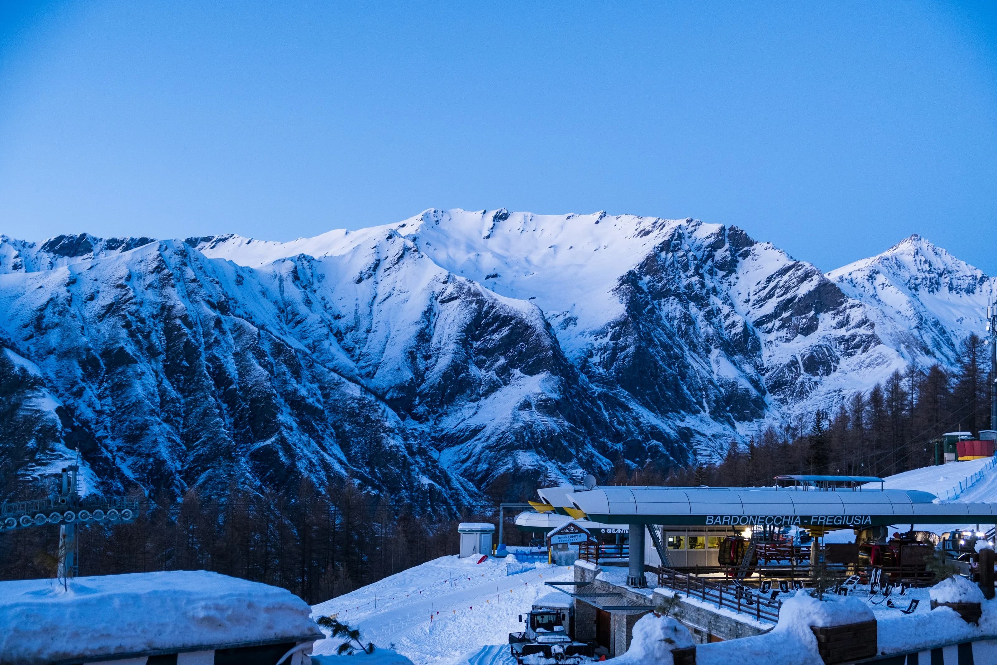 Bardonecchia d’inverno all’ora blu: le montagne innevate sopra la stazione degli impianti Bardonecchia-Fregiusia e una pista battuta