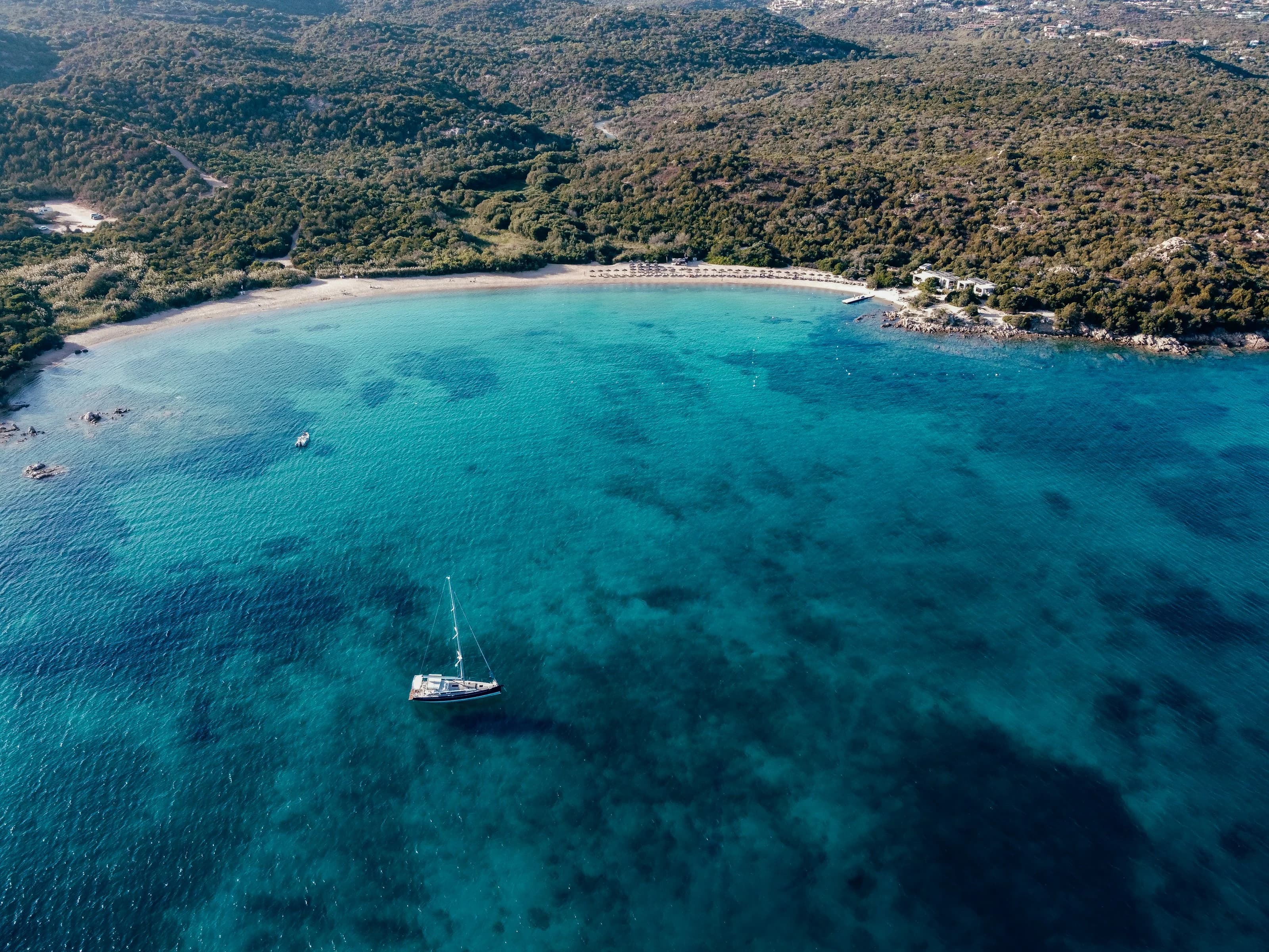 The bay of Cala di Volpe from above: turquoise water, a sailing boat at anchor and a pale beach against the Mediterranean scrub