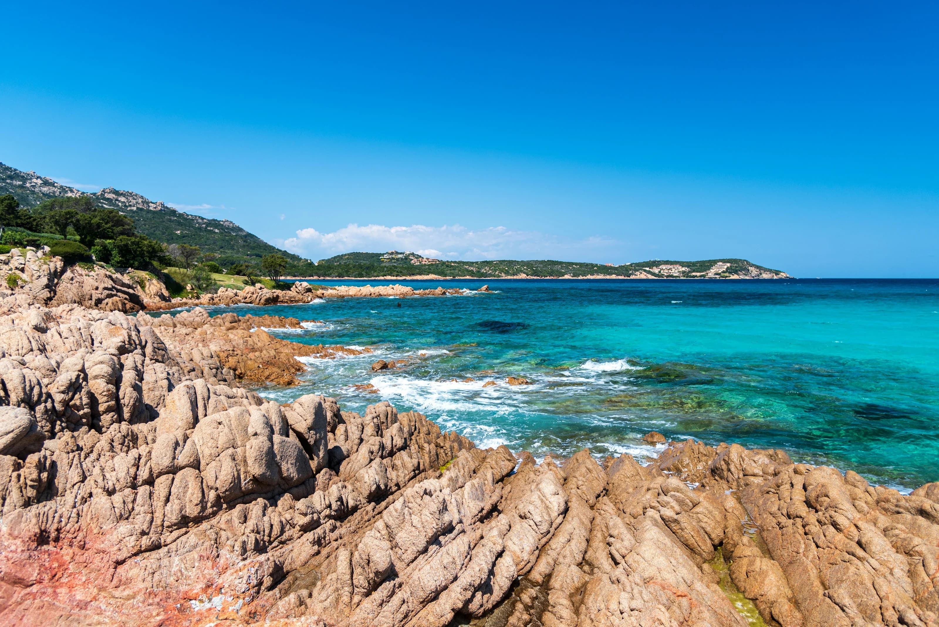 Scogli di granito rosato in primo piano e il mare turchese del Grande Pevero, con le colline della costa sullo sfondo
