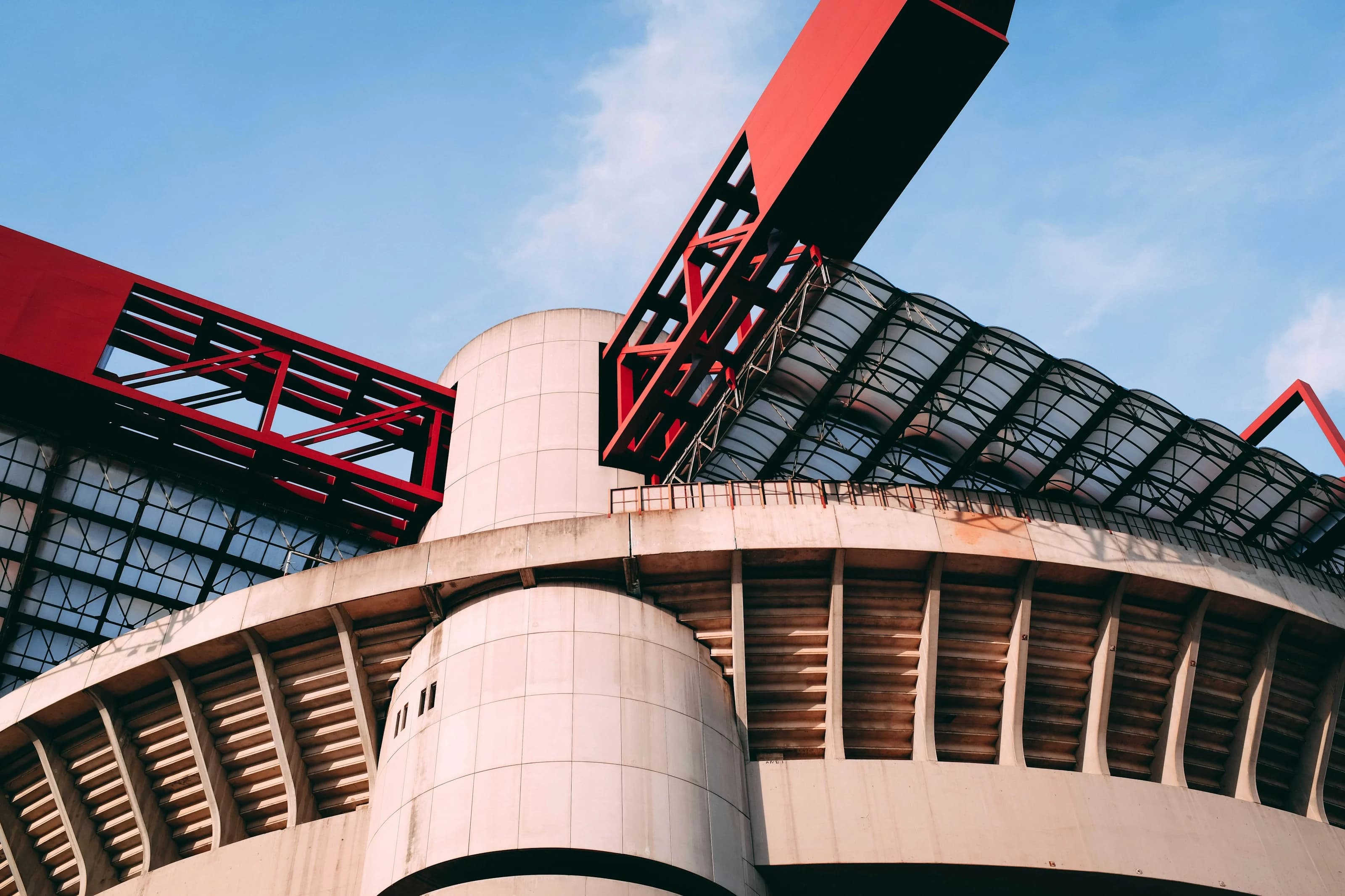 Lo stadio di San Siro visto dal basso: le torri cilindriche di cemento, le rampe a spirale e le travi rosse della copertura contro il cielo azzurro