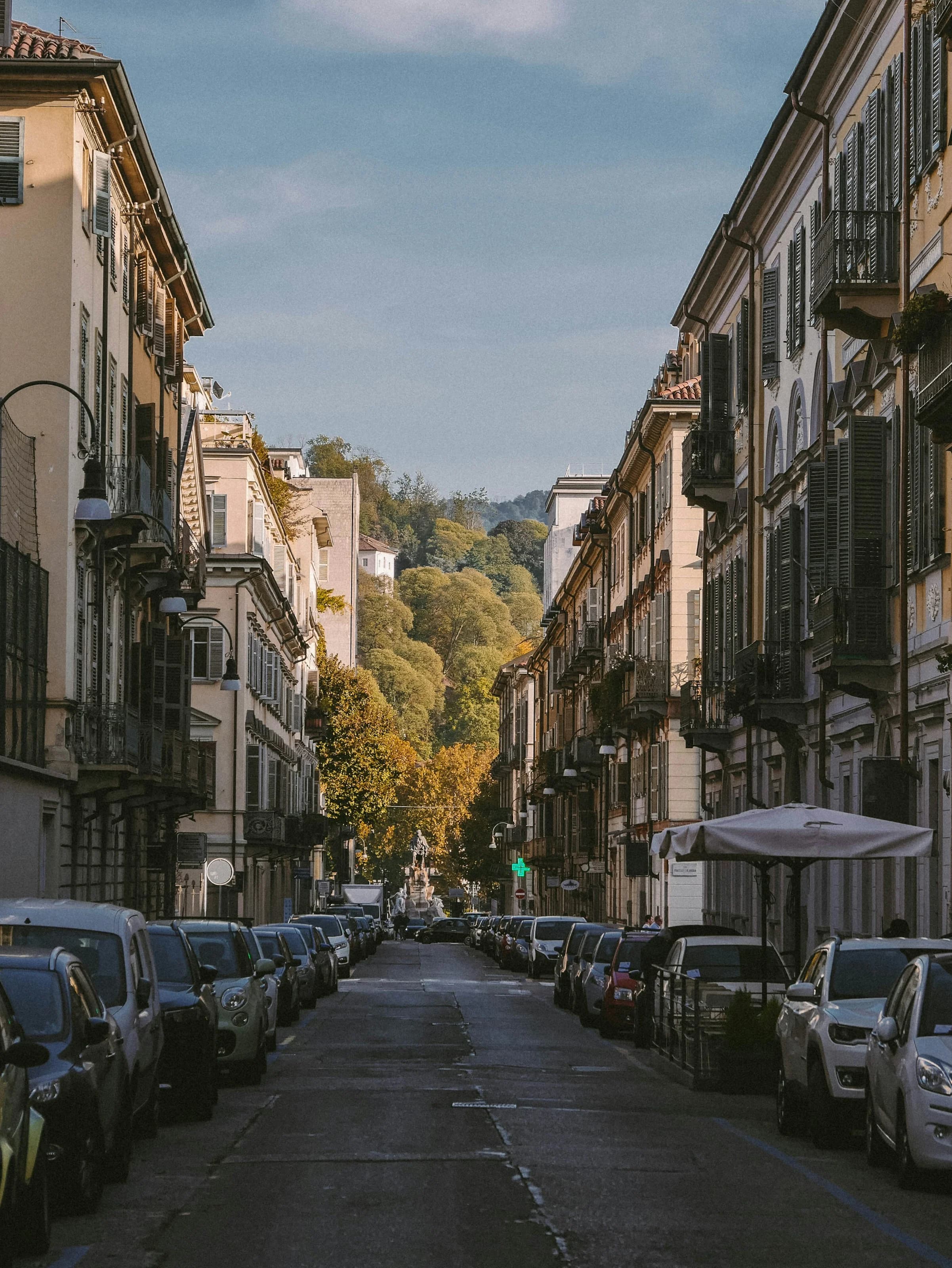 Una via residenziale di Torino con palazzi a balconi e persiane, le auto parcheggiate sui due lati e, in fondo, gli alberi della collina in autunno