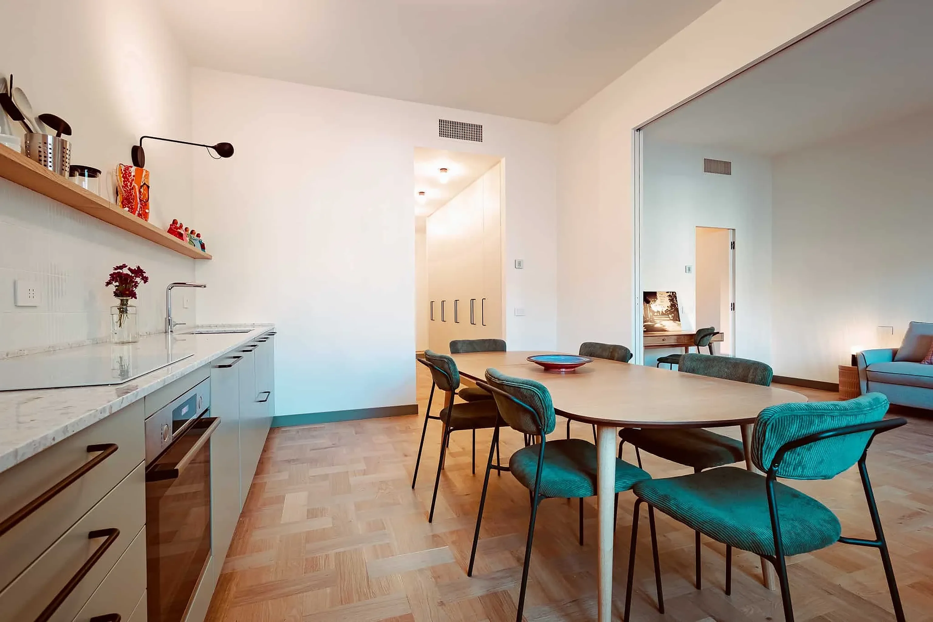 A kitchen with a pale marble worktop and an oval wooden table with green chairs; beyond a sliding door, the living room