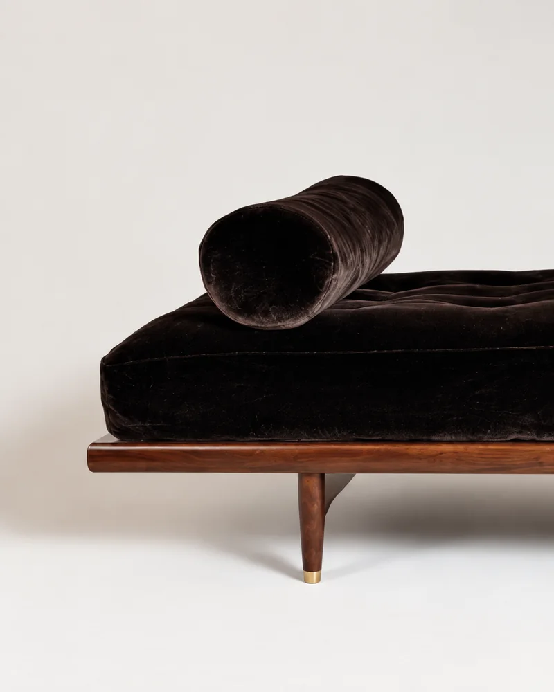 A dark brown velvet daybed with a bolster cushion and a walnut frame