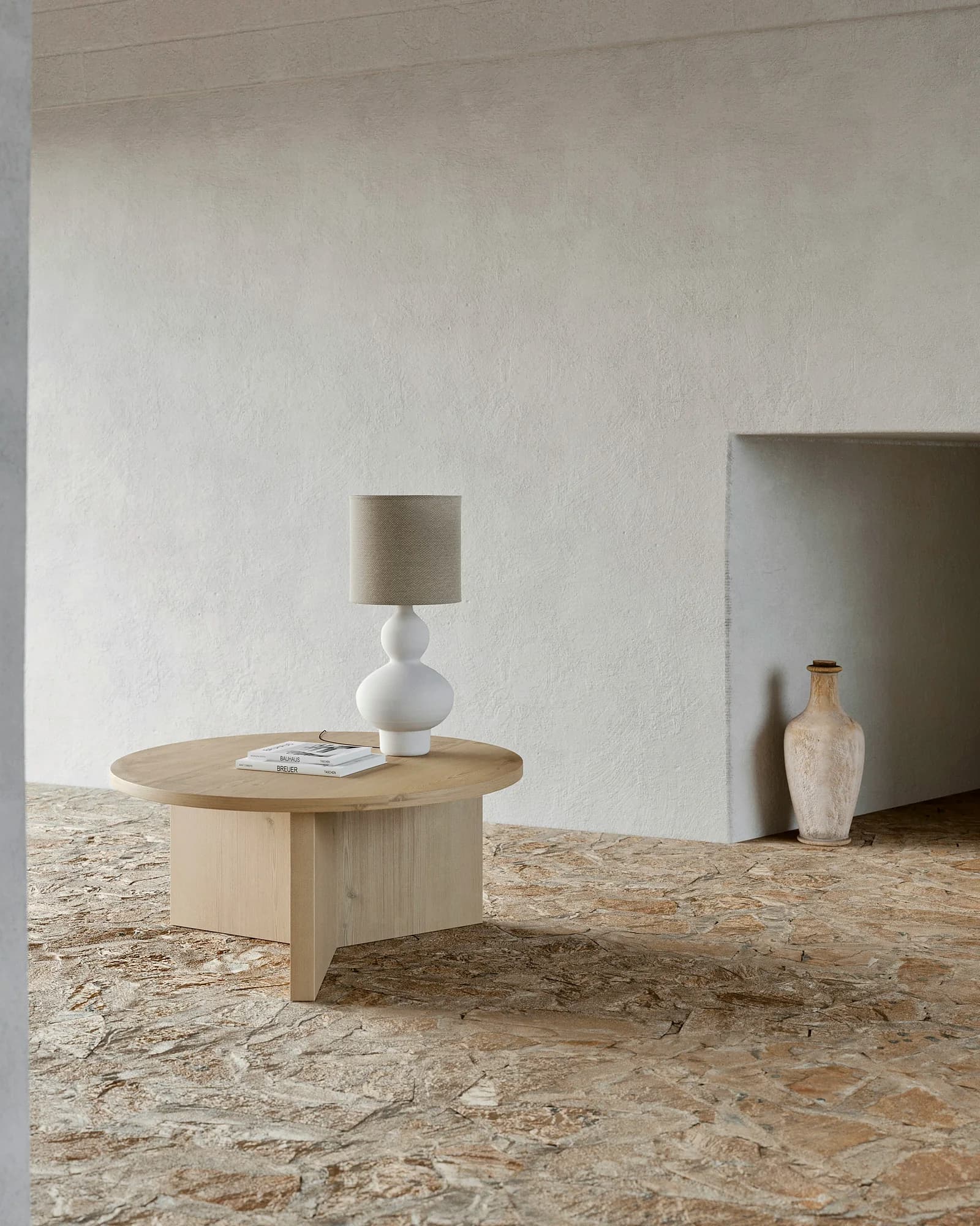 A bare travertine room with a round stone coffee table, a lamp and a vase