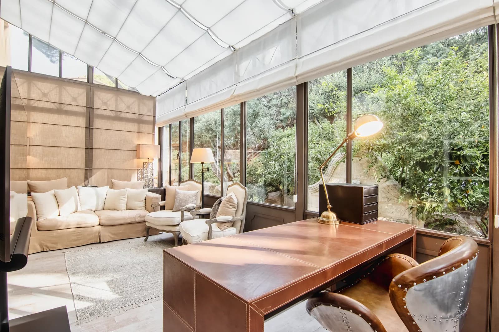A glass veranda with a leather-covered desk and a lit brass lamp; behind it, a pale sofa, two armchairs and the garden beyond the glass
