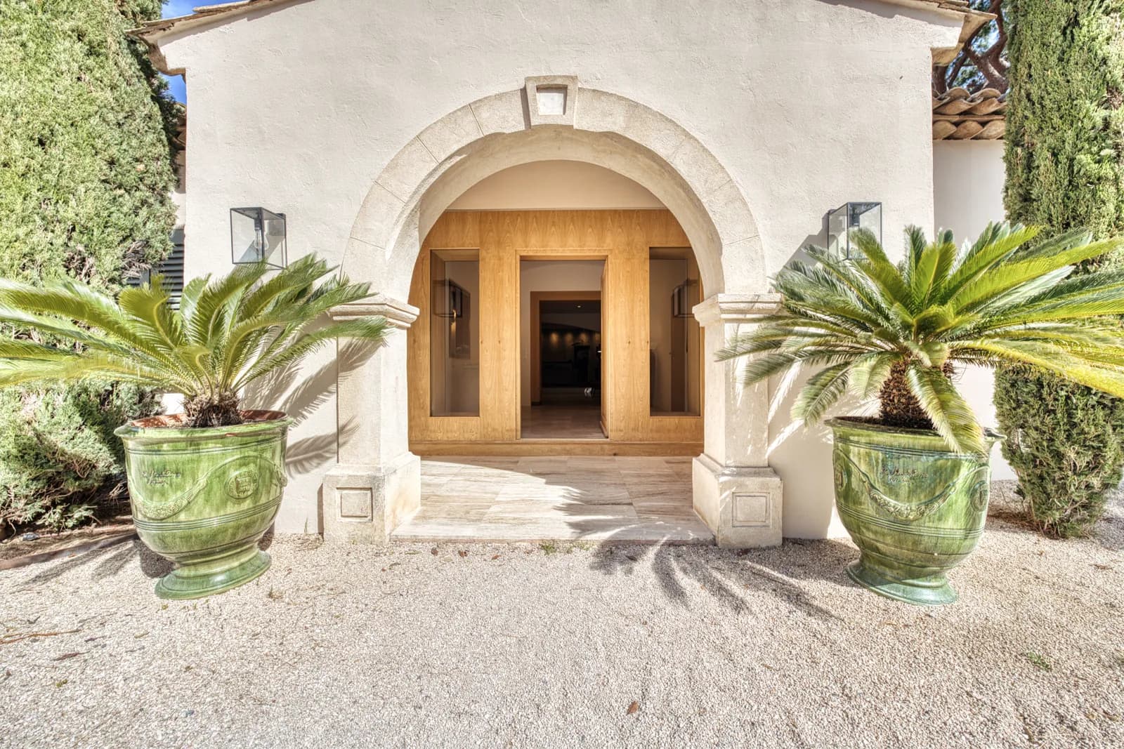 Un ingresso ad arco in pietra chiara con la porta di legno aperta, fra due grandi vasi verdi con palme nane e due lanterne alle pareti